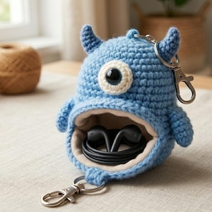 May include: A handmade blue crochet monster-shaped pouch with a large open mouth, holding black earbuds. The monster has one large eye and small horns. It includes a silver keychain clip. Yarn and crochet hooks are in the background.