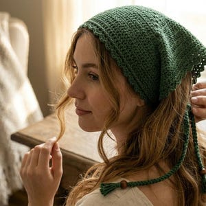 Puede incluir: Un gorro verde tejido a ganchillo con forma cuadrada y borde festoneado. El gorro tiene largas cintas con cuentas de madera. El gorro lo lleva una persona con pelo largo y ondulado.