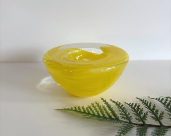 May include: A yellow glass bowl with a white swirl design. The bowl has a rounded shape and sits on a white surface. A green fern leaf is in the foreground. The bowl is a decorative item.