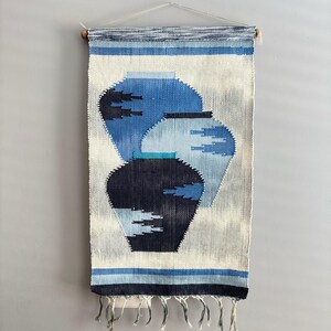 May include: Woven wall hanging featuring three stylized blue vases against a white and gray background. The vases have varying shades of blue and are outlined with a darker blue. The top has a striped design, and the bottom has fringe.