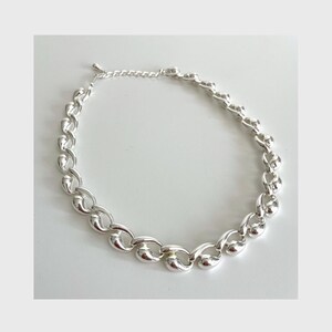 May include: A silver-colored necklace with a chunky chain design. The necklace features large, rounded links that create a bold and modern look. The chain is connected with a delicate chain and clasp.