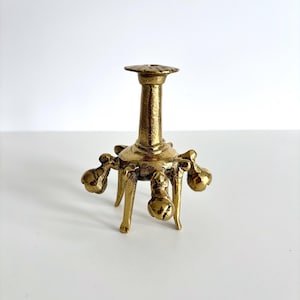 May include: A gold-colored metal candle holder with a round top and a long, slender stem. The base has three legs and decorative bell-shaped accents. The candle holder is designed to hold a single candle.