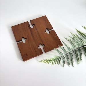May include: A square, interlocking wooden trivet with a warm brown finish. The trivet is made of four pieces that fit together, with a cross-shaped cutout on each side. The trivet has the text "DANSK" and "DENMARK" printed on one of the pieces.