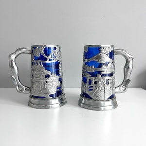 May include: Two decorative beer steins with cobalt blue glass interiors. The silver-colored metal exteriors feature detailed relief carvings of Asian-inspired scenes. Each stein has a handle with a dragon-like design.