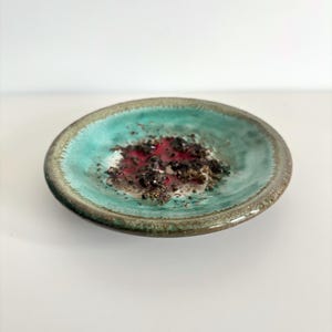 May include: A small, round ceramic dish with a turquoise interior and a dark brown rim. The center of the dish contains a mix of red, black, and gold-colored materials, creating a textured appearance. The dish is set against a white background.