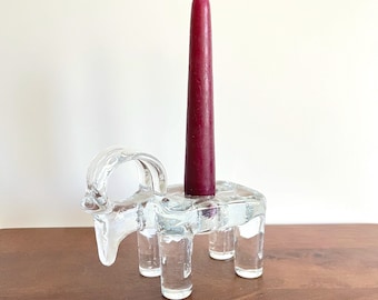 May include: A clear glass candle holder shaped like a stylized animal with four legs. A burgundy candle is placed in the holder.