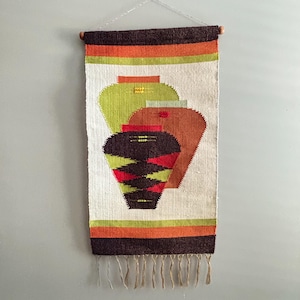 May include: Woven wall hanging featuring three stylized vase shapes in shades of green, brown, and red against a cream background. The top and bottom have horizontal stripes in brown, orange, and green. The bottom edge has fringe.
