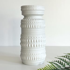 May include: A white ceramic vase with a textured surface. The vase has a cylindrical shape with various embossed patterns, including dots, diamonds, and horizontal lines. The vase is approximately 10 inches tall.