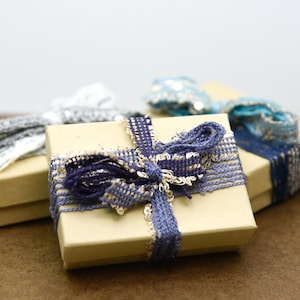 Puede incluir: Tres peque&ntilde;as cajas de regalo envueltas en papel marr&oacute;n con cintas de diferentes colores. Las cajas est&aacute;n atadas con cintas azules, grises y blancas.