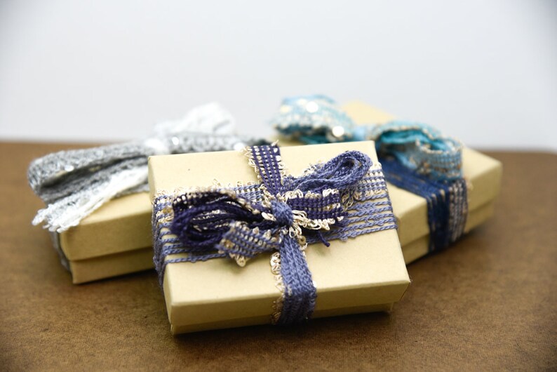Puede incluir: Tres peque&ntilde;as cajas de regalo envueltas en papel marr&oacute;n con cintas de diferentes colores. Las cajas est&aacute;n atadas con cintas azules, grises y blancas.