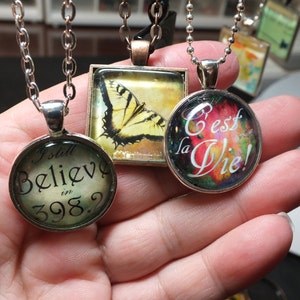 Puede incluir: Tres collares de plata con colgantes de vidrio. Los colgantes presentan una mariposa, un dise&ntilde;o abstracto colorido con el texto "C'est la Vie!" y un dise&ntilde;o de estilo vintage con el texto "I still believe in 398?"