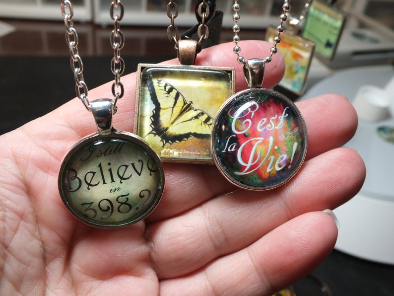 Puede incluir: Tres collares de plata con colgantes de vidrio. Los colgantes presentan una mariposa, un dise&ntilde;o abstracto colorido con el texto "C'est la Vie!" y un dise&ntilde;o de estilo vintage con el texto "I still believe in 398?"