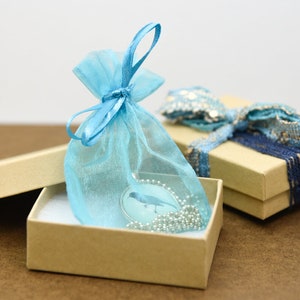 Puede incluir: Un collar con un colgante de p&aacute;jaro azul en una bolsa de regalo de organza azul claro, dentro de una caja de regalo marr&oacute;n. La caja est&aacute; abierta y la bolsa est&aacute; sentada encima del papel de seda blanco dentro de la caja.