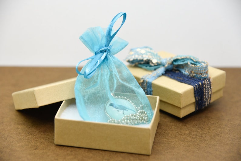 Puede incluir: Un collar con un colgante de p&aacute;jaro azul en una bolsa de regalo de organza azul claro, dentro de una caja de regalo marr&oacute;n. La caja est&aacute; abierta y la bolsa est&aacute; sentada encima del papel de seda blanco dentro de la caja.