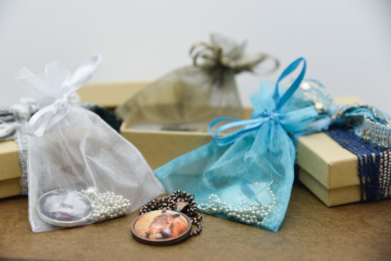 Puede incluir: Dos peque&ntilde;as bolsas de regalo, una blanca y otra azul, con cuentas plateadas y blancas y un collar con un colgante redondo. Las bolsas est&aacute;n sobre una superficie marr&oacute;n.