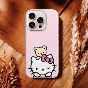 Puede incluir: Funda de tel&eacute;fono rosa con un dise&ntilde;o de dibujos animados de Hello Kitty y un oso de peluche. La funda tiene un borde transparente y un recorte para la c&aacute;mara. El fondo es una superficie de madera con plantas secas.