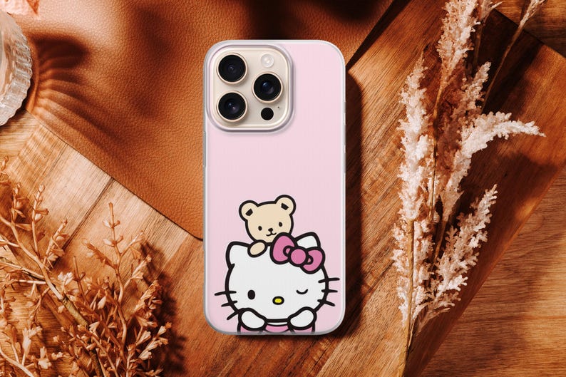 Puede incluir: Funda de tel&eacute;fono rosa con un dise&ntilde;o de dibujos animados de Hello Kitty y un oso de peluche. La funda tiene un borde transparente y un recorte para la c&aacute;mara. El fondo es una superficie de madera con plantas secas.