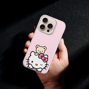 Puede incluir: Funda de tel&eacute;fono rosa con un dise&ntilde;o de Hello Kitty y un peque&ntilde;o oso de peluche. La funda tiene un borde transparente y un recorte para la c&aacute;mara. El dise&ntilde;o incluye un lazo rosa y contornos negros. La funda del tel&eacute;fono se sostiene en una mano.