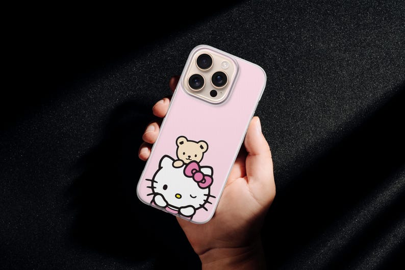 Puede incluir: Funda de tel&eacute;fono rosa con un dise&ntilde;o de Hello Kitty y un peque&ntilde;o oso de peluche. La funda tiene un borde transparente y un recorte para la c&aacute;mara. El dise&ntilde;o incluye un lazo rosa y contornos negros. La funda del tel&eacute;fono se sostiene en una mano.