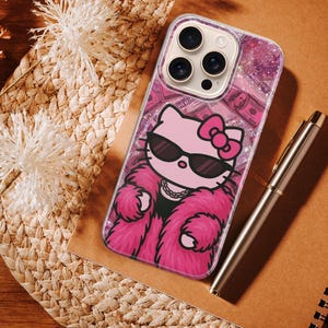 Puede incluir: Una funda transparente para tel&eacute;fono con un dise&ntilde;o de Hello Kitty rosa que lleva gafas de sol y un abrigo de piel rosa. La funda tiene un fondo brillante con billetes de d&oacute;lar. Un bol&iacute;grafo plateado y un cuaderno de espiral tambi&eacute;n est&aacute;n en la imagen.