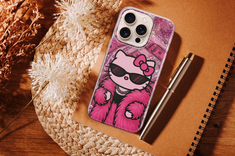 Puede incluir: Una funda transparente para tel&eacute;fono con un dise&ntilde;o de Hello Kitty rosa que lleva gafas de sol y un abrigo de piel rosa. La funda tiene un fondo brillante con billetes de d&oacute;lar. Un bol&iacute;grafo plateado y un cuaderno de espiral tambi&eacute;n est&aacute;n en la imagen.