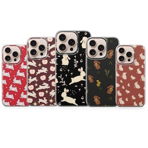 Puede incluir: Cinco fundas para smartphone dispuestas en fila, cada una con un patrón distinto de conejos. Los diseños incluyen fondos rojos, rojo oscuro, negros y marrones con varias ilustraciones de conejos blancos o marrones, manzanas y flores.