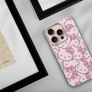 Puede incluir: Una funda de tel&eacute;fono rosa con un patr&oacute;n repetido de caras de Hello Kitty y lazos rosas. La funda est&aacute; sobre una superficie gris claro, cerca de una obra de arte enmarcada con un marco negro.