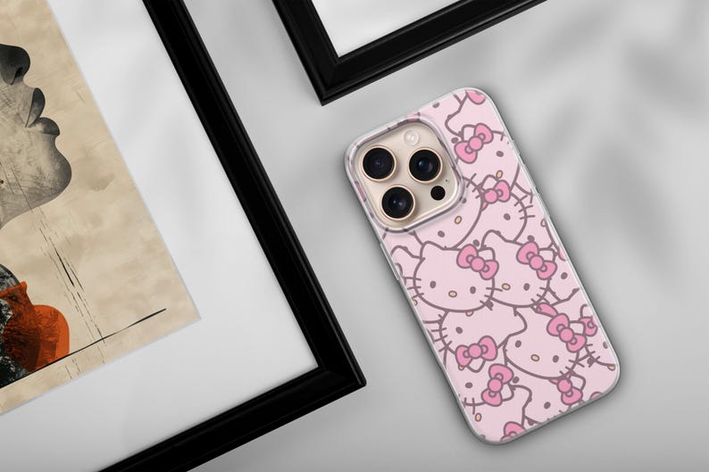 Puede incluir: Una funda de tel&eacute;fono rosa con un patr&oacute;n repetido de caras de Hello Kitty y lazos rosas. La funda est&aacute; sobre una superficie gris claro, cerca de una obra de arte enmarcada con un marco negro.