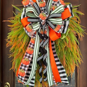 May include: A decorative wreath featuring a large bow made of ribbons with orange pumpkin prints, black and white buffalo check patterns, and green and white stripes. The base consists of cascading green and orange fern-like foliage.
