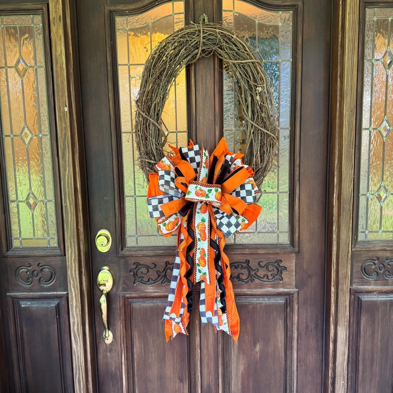 May include: A circular grapevine wreath featuring a large decorative bow at the bottom. The bow is constructed from ribbons in solid orange, black and white checkered patterns, and a white ribbon printed with small orange pumpkins and green leaves.