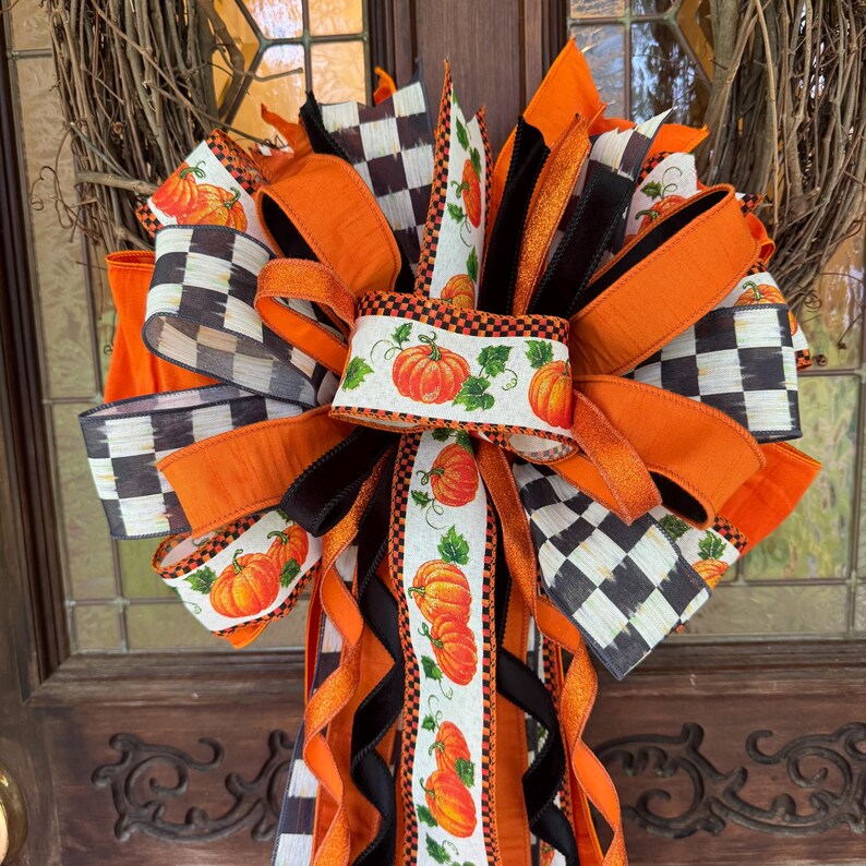 May include: A decorative wreath bow featuring a mix of orange, black, and white ribbons. The design includes solid orange ribbons, black and white checkered patterns, and white ribbons printed with orange pumpkins and green leaves.