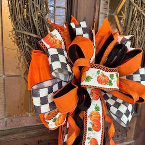 May include: A decorative wreath bow featuring a mix of solid orange ribbons, black and white checkered ribbons, and white ribbons with a pumpkin and green leaf print. The bow is constructed from multiple loops and long tails of wired ribbon.