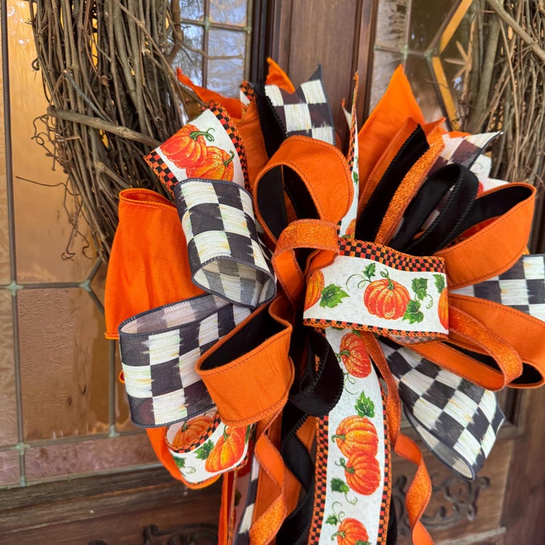 May include: A decorative wreath bow featuring a mix of solid orange ribbons, black and white checkered ribbons, and white ribbons with a pumpkin and green leaf print. The bow is constructed from multiple loops and long tails of wired ribbon.