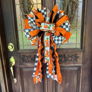 May include: A decorative wreath featuring a natural twig base adorned with a large, multi-layered bow. The bow is crafted from ribbons in solid orange, black, and white checkered patterns, and a white ribbon printed with orange pumpkin motifs.
