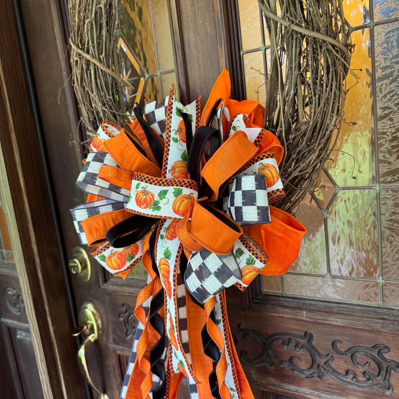 May include: A decorative wreath made of brown twigs featuring a large, multi-layered bow. The bow is constructed from ribbons in solid orange, black, and white checkered patterns, and a white ribbon printed with orange pumpkins and green vines.