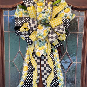 May include: A decorative bow made of wired ribbon featuring a lemon pattern, black and white polka dots, black and white buffalo check, and yellow gingham. The bow has multiple loops and long, flowing tails with wired edges for shaping.