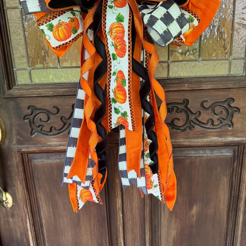 May include: A decorative bow featuring multiple ribbons in orange, black, and white. The ribbons include patterns of orange pumpkins with green leaves and black and white checkered designs. The bow has long, flowing tails with varying textures.