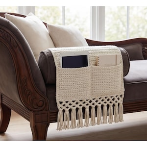 May include: Cream-colored, crocheted couch armrest organizer with two pockets and a fringe. A tablet and a notebook are visible in the pockets. The organizer is draped over a brown leather couch with decorative wooden armrests.