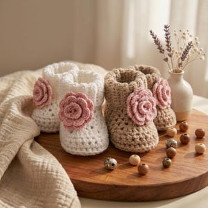 May include: Four handmade crochet baby booties. Two are white, and two are tan. Each bootie has a pink flower accent. The booties are displayed on a wooden tray with decorative beads and a small vase with flowers.