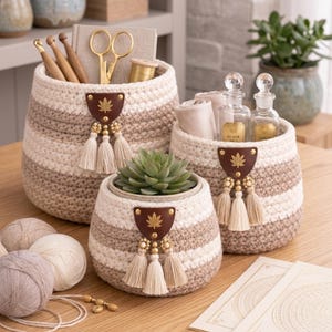 May include: Set of three handmade crochet baskets in cream and brown stripes. Each basket features a brown leather tag with a leaf design, adorned with tassels and beads. The baskets are filled with craft supplies and a succulent.