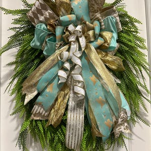 Puede incluir: Una corona decorativa con una base de follaje verde, adornada con múltiples capas de cintas. Las cintas incluyen oro, blanco y turquesa con patrones de estrellas doradas y acentos marrones estampados. Para la decoración del hogar.