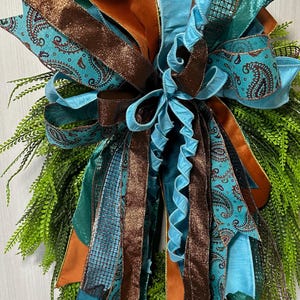 May include: A decorative wreath featuring a large bow with multiple ribbons in teal, brown, and orange. The ribbons have paisley and glitter patterns. The wreath has a base of green foliage.