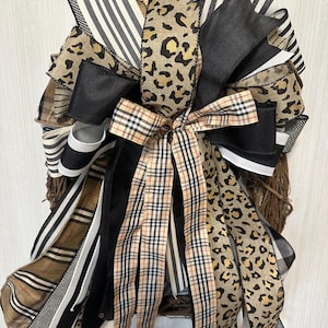 May include: A decorative wreath featuring a variety of ribbons in black, white, and tan. The ribbons have patterns including stripes, leopard print, and plaid. The wreath is made of woven branches.