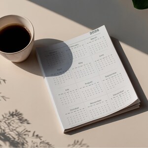 Op de afbeelding: Een kalender voor 2026 met maandelijkse lay-outs, naast een kopje koffie. De kalender is open op een pagina met de maanden van het jaar. De beker is wit en de koffie is donker. De scène wordt verlicht door natuurlijk licht.