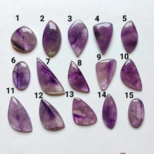 May include: Fifteen polished amethyst cabochons arranged in three rows, numbered 1 through 15. Each stone displays varying shades of purple and white banding in diverse shapes, including oval, teardrop, triangular, and irregular geometric forms.