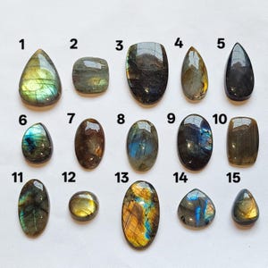 May include: A collection of fifteen polished labradorite gemstones arranged in three rows, numbered one through fifteen. Each stone displays unique iridescent flashes of blue, gold, and green against a dark gray base, featuring various shapes including teardrop, oval, and rectangular cuts.