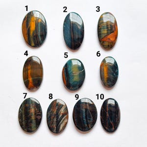 May include: Ten oval-shaped polished gemstones arranged in rows, numbered one through ten. Each stone features unique chatoyant patterns in shades of deep blue, golden brown, and black, with a smooth, reflective surface and distinct color banding.