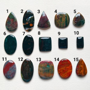 May include: A collection of fifteen polished gemstones arranged in three rows, numbered one through fifteen. The stones feature various shapes including teardrop, oval, rectangular, and square, displaying diverse colors like deep green, red, and brown.