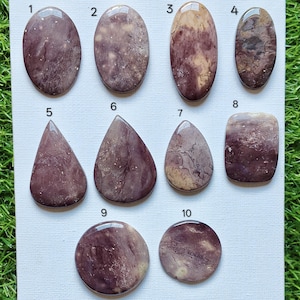 May include: Ten polished cabochon gemstones arranged on a white surface, numbered one through ten. These stones feature varied shapes including ovals, teardrops, a rectangle, and circles, displaying mottled patterns in shades of plum, tan, and cream.