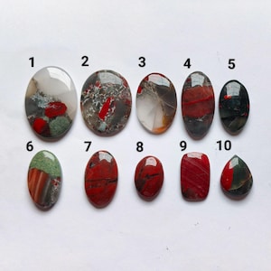 May include: Ten polished cabochon gemstones arranged in two rows, numbered one through ten. The stones feature varied patterns and colors including red, green, gray, and white, with oval, rectangular, and teardrop shapes displayed on a white background.